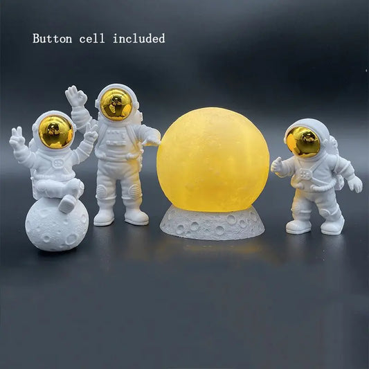 The Astronaut Yellow Moon Ambient Light Ornament by HomeForDecor features three astronaut figurines with gold visors arranged around a glowing yellow moon on a gray base. One of the astronauts sits atop a miniature lunar surface, adding to the charm of this ornament. The text notes that a button cell is included, and the dark, neutral background amplifies the ambient light effect.