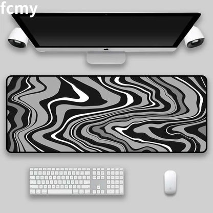 Under an Apple keyboard, mouse, monitor, and speakers on a gray desk lays the Art Strata Liquid Mouse Pad by HomeForDecor, featuring a sleek grayscale abstract wavy design. It is expertly crafted with durable fabric through the sublimation process.