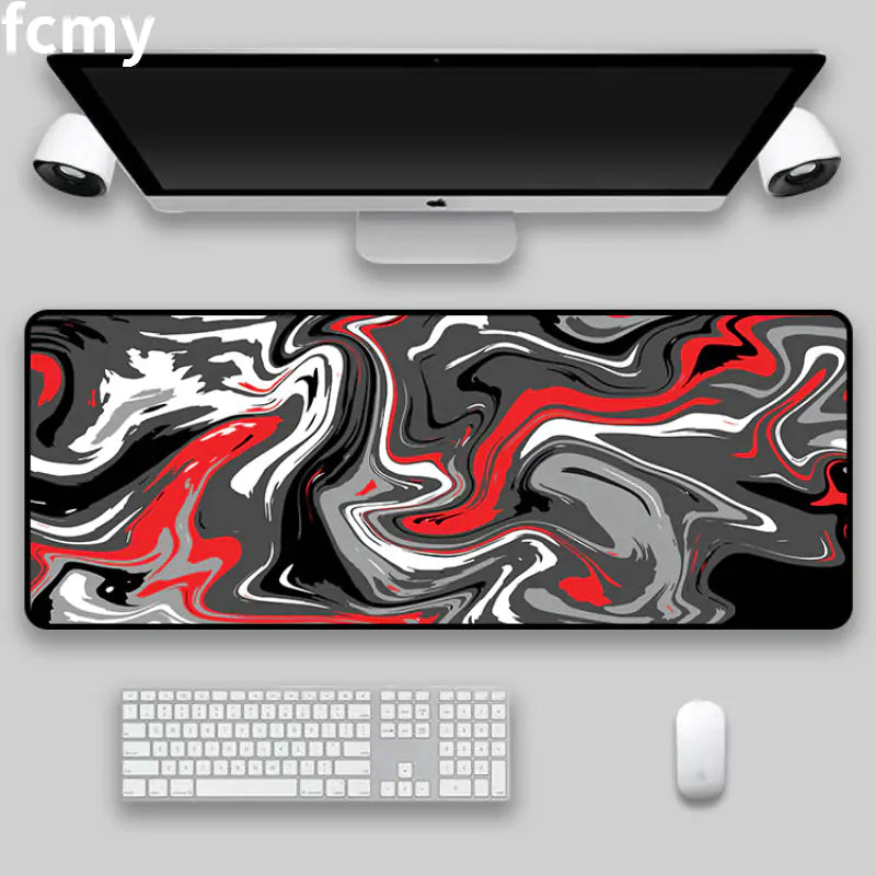 A computer desk setup featuring the Art Strata Liquid Mouse Pad from HomeForDecor, characterized by a large, abstract swirl pattern in black, white, and red on durable fabric. The sleek monitor is accompanied by two wall-mounted speakers, a keyboard, and a mouse arranged neatly on the desk.